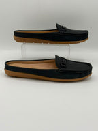 A pair of black leather mule loafers with tan soles, displayed on a white studio background with one shoe resting on a clear acrylic riser and the other lying flat.