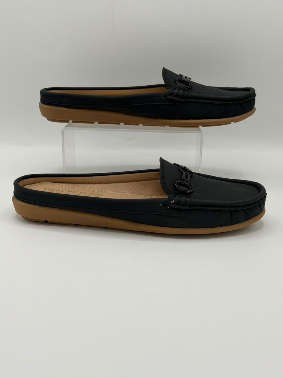 A pair of black leather mule loafers with tan soles, displayed on a white studio background with one shoe resting on a clear acrylic riser and the other lying flat.