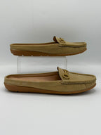 A pair of tan slip-on mule loafers displayed on a white studio background, with one shoe resting flat and the other elevated on a clear acrylic riser.