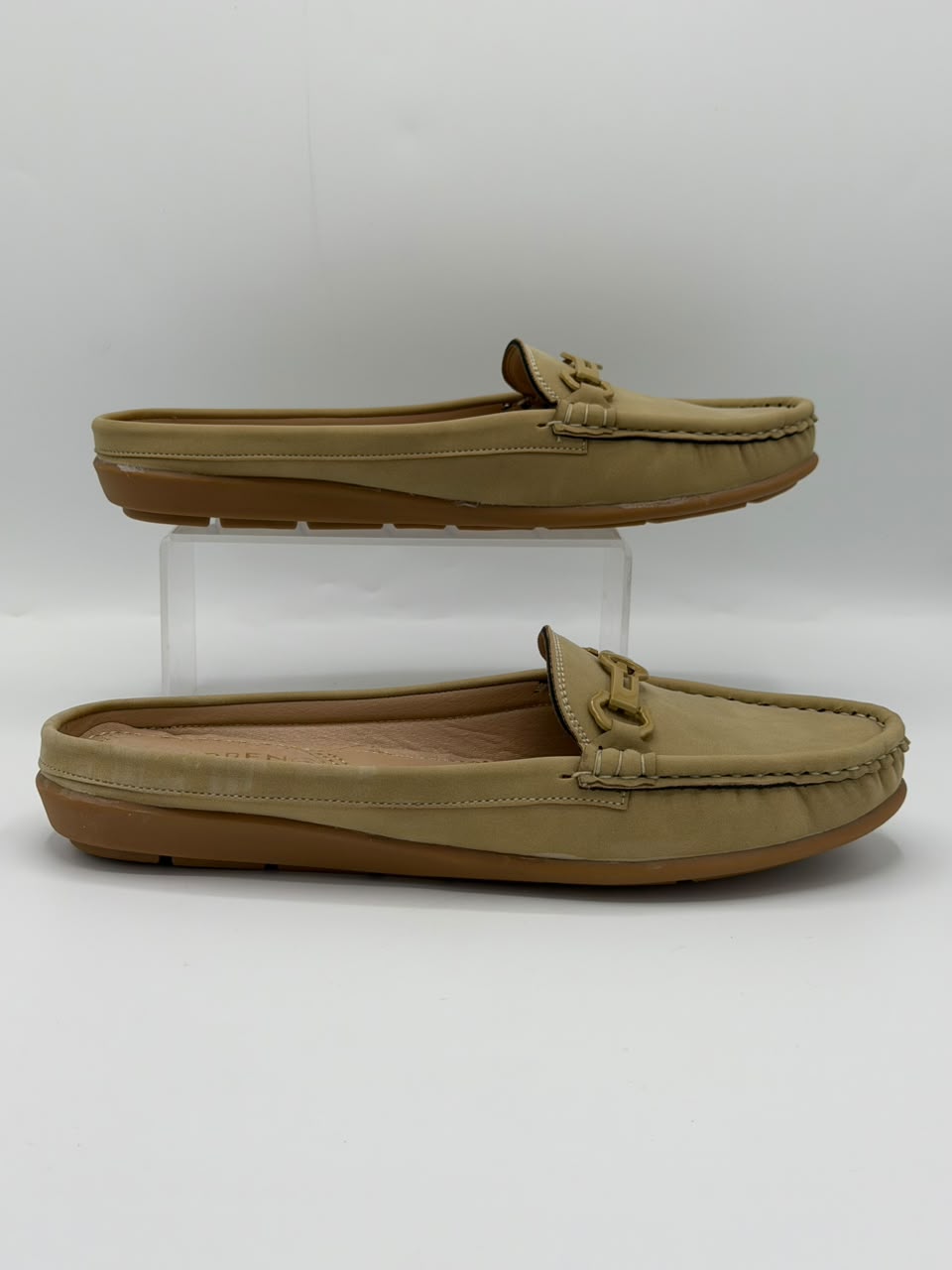 A pair of tan slip-on mule loafers displayed on a white studio background, with one shoe resting flat and the other elevated on a clear acrylic riser.