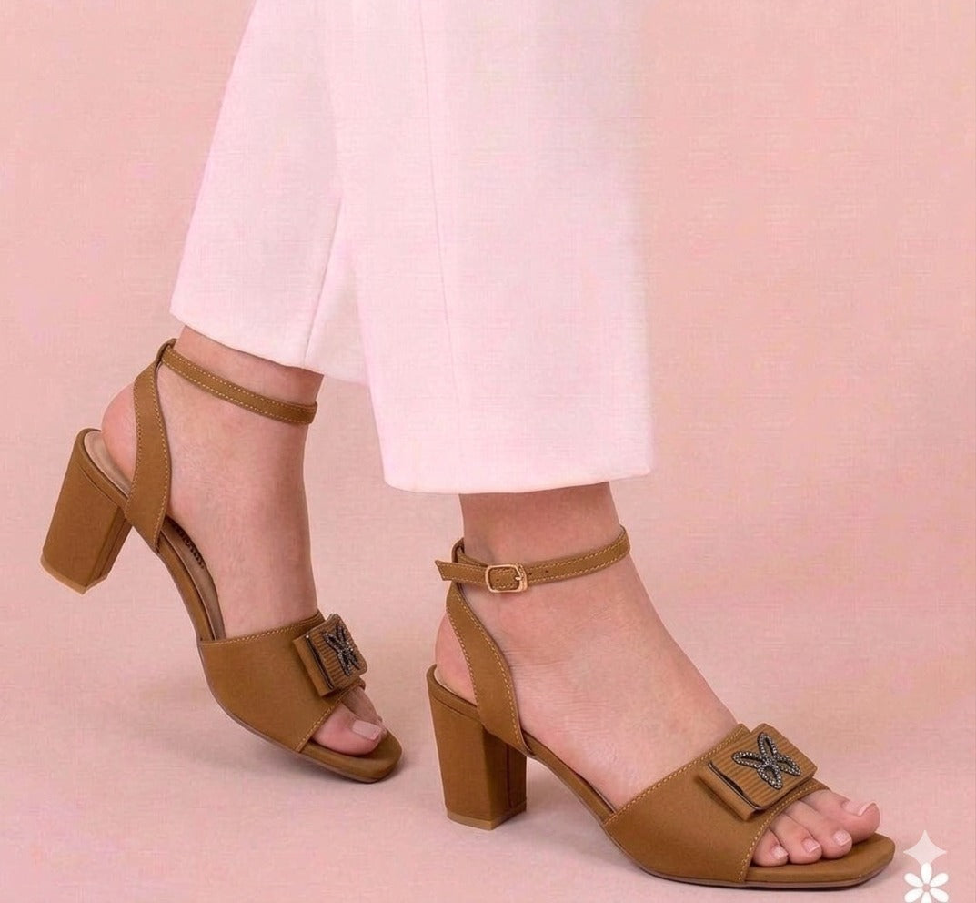 Brown block-heel sandals with decorative buckles worn by a person on a pink background