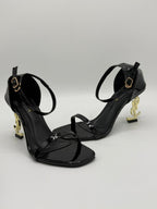 Black high-heeled sandals with gold accents on a light gray background