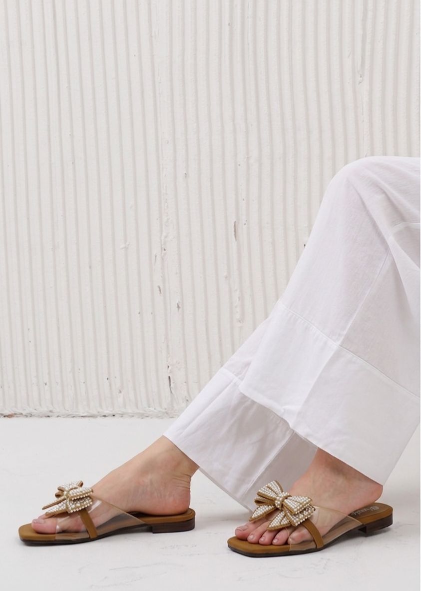 Brown sandals with bow details worn with white pants against a light wooden panel background