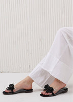 Black sandals with bow details worn with white pants against a light background