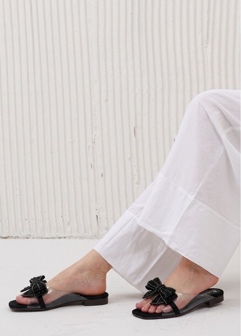 Black sandals with bow details worn with white pants against a light background