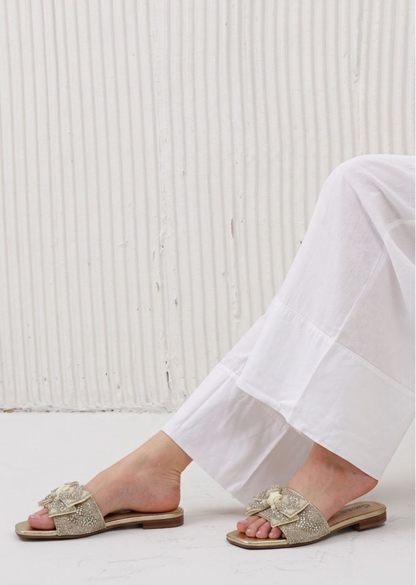 Gold sandals worn with white pants against a light wooden floor background