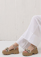 Brown platform sandals with floral details worn by a person in white pants against a light wooden panel background.