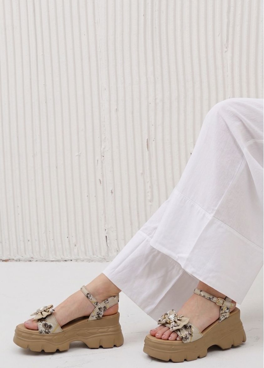 Brown platform sandals with floral details worn by a person in white pants against a light wooden panel background.