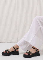 Black sandals with floral straps worn by a person sitting on a white floor against a white wall.