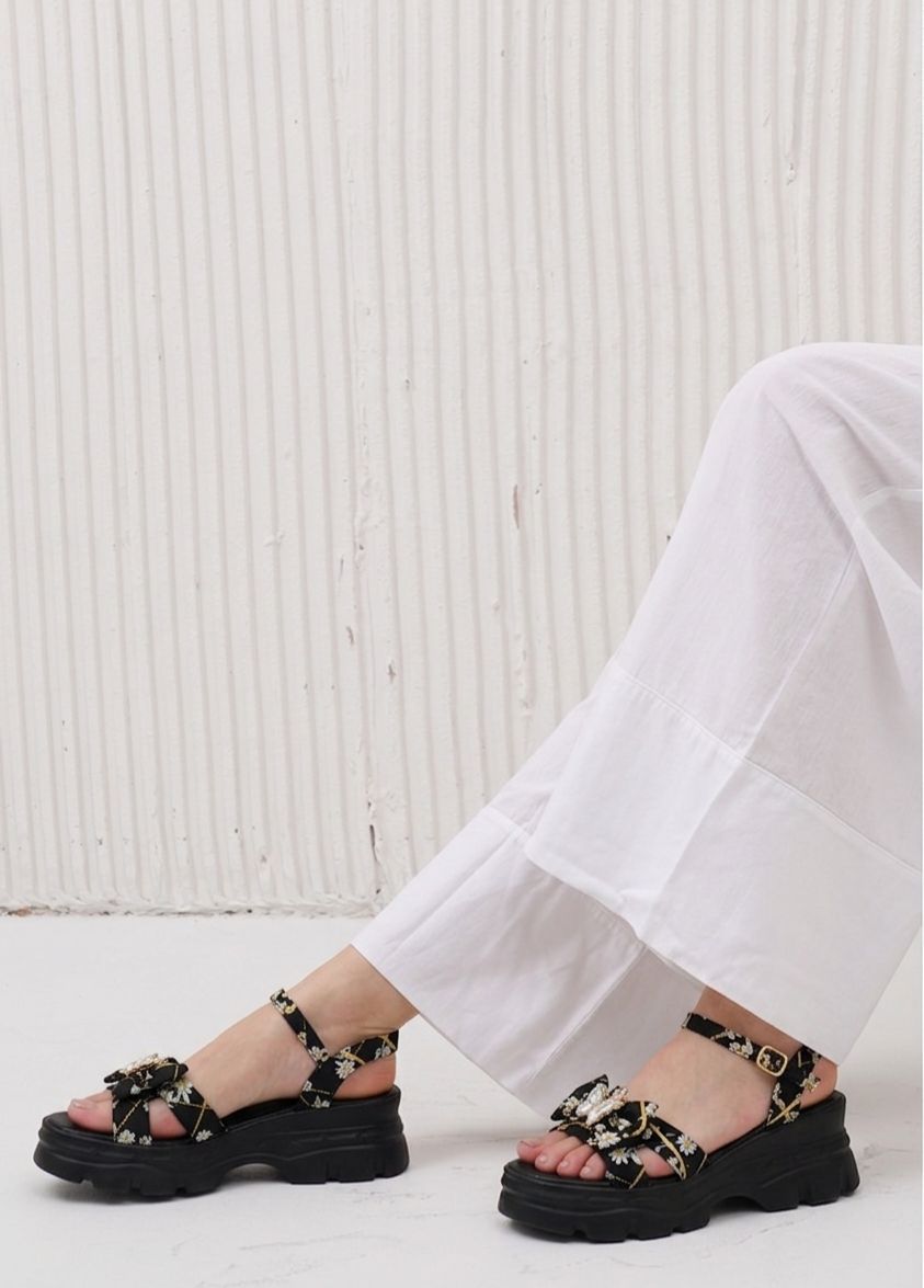 Black sandals with floral straps worn by a person sitting on a white floor against a white wall.