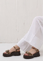Brown sandals with decorative straps worn by a person in white pants against a light background