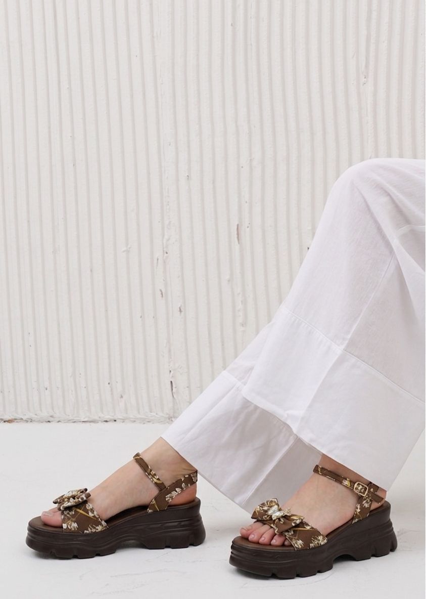 Brown sandals with decorative straps worn by a person in white pants against a light background
