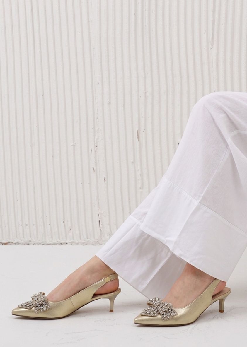 Gold high-heeled shoes with embellishments worn with white pants against a white background