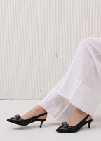 Black high-heeled shoes with decorative elements worn with white pants on a light background