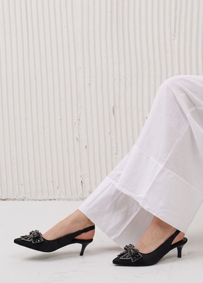 Black high-heeled shoes with decorative elements worn with white pants on a light background
