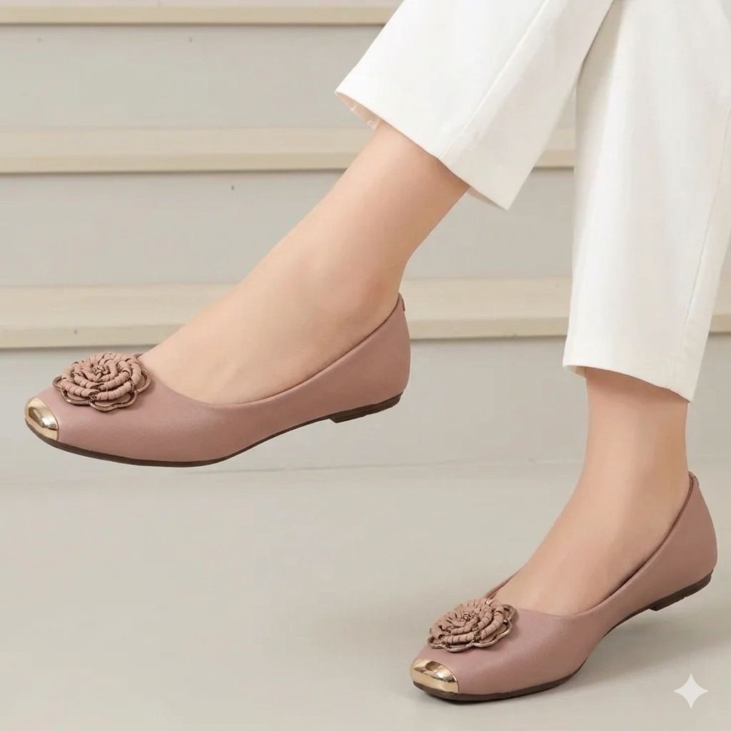 Pink flat shoes with floral embellishments worn by a person in white pants on a neutral background
