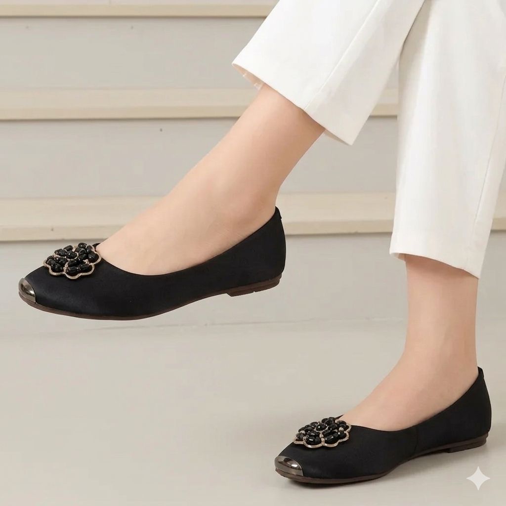 Black flat shoes with floral embellishments worn by a person in white pants on a light background